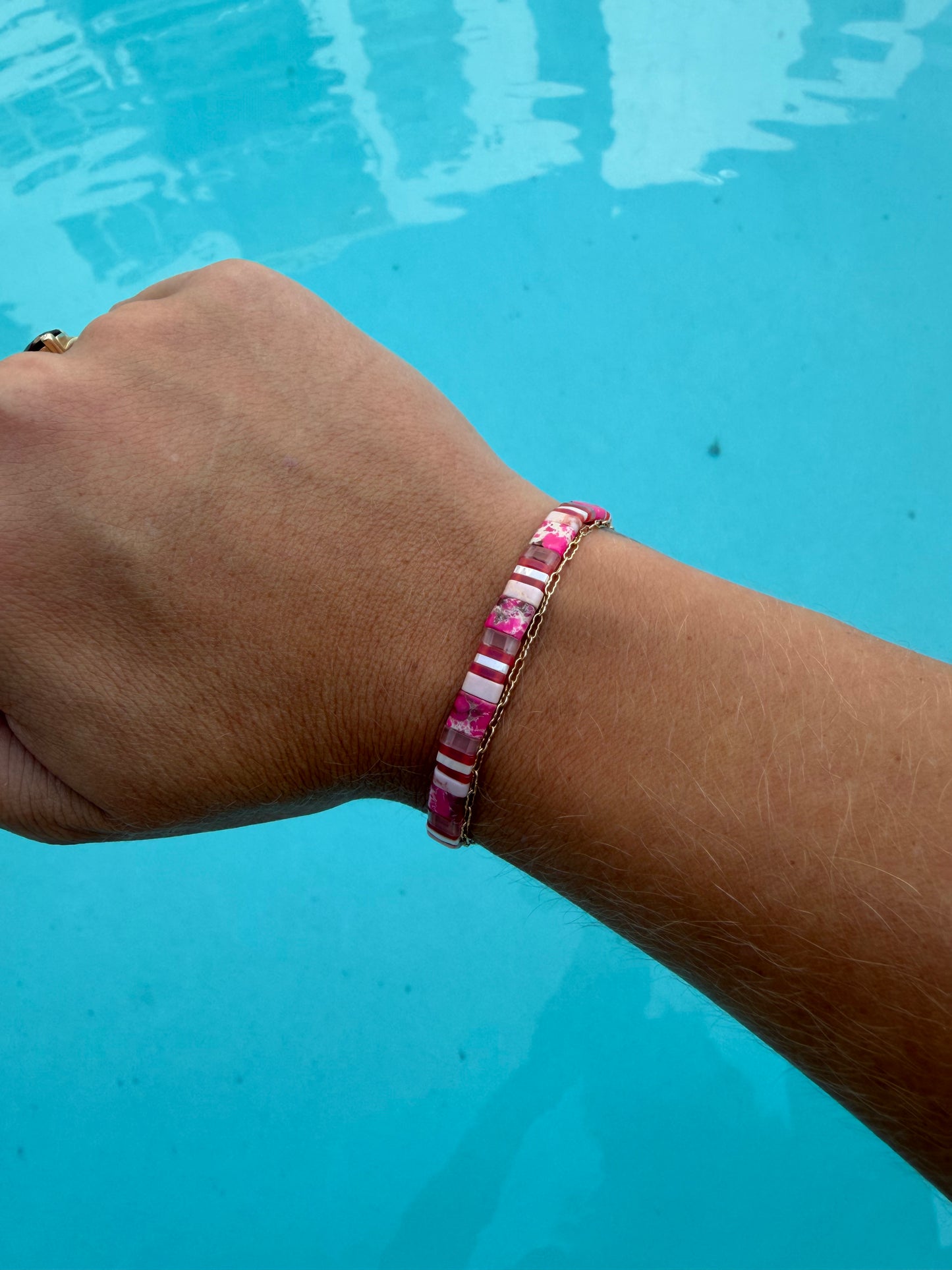 Handmade Pink Sea Star bracelet worn on a wrist beside a gold chain bracelet over blue pool water.