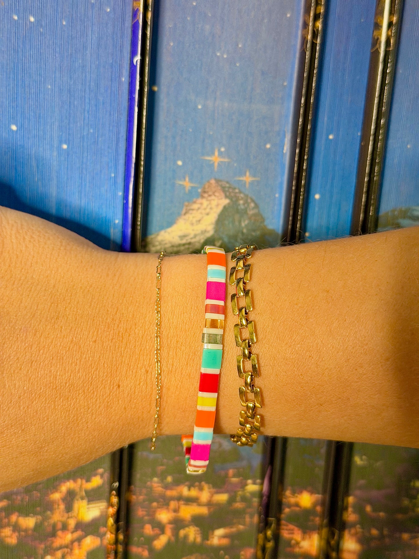 Fantasy Bookshelf Bracelet Second Edition stacked with two gold bracelets on a wrist against blue book spines featuring mountain and star artwork.