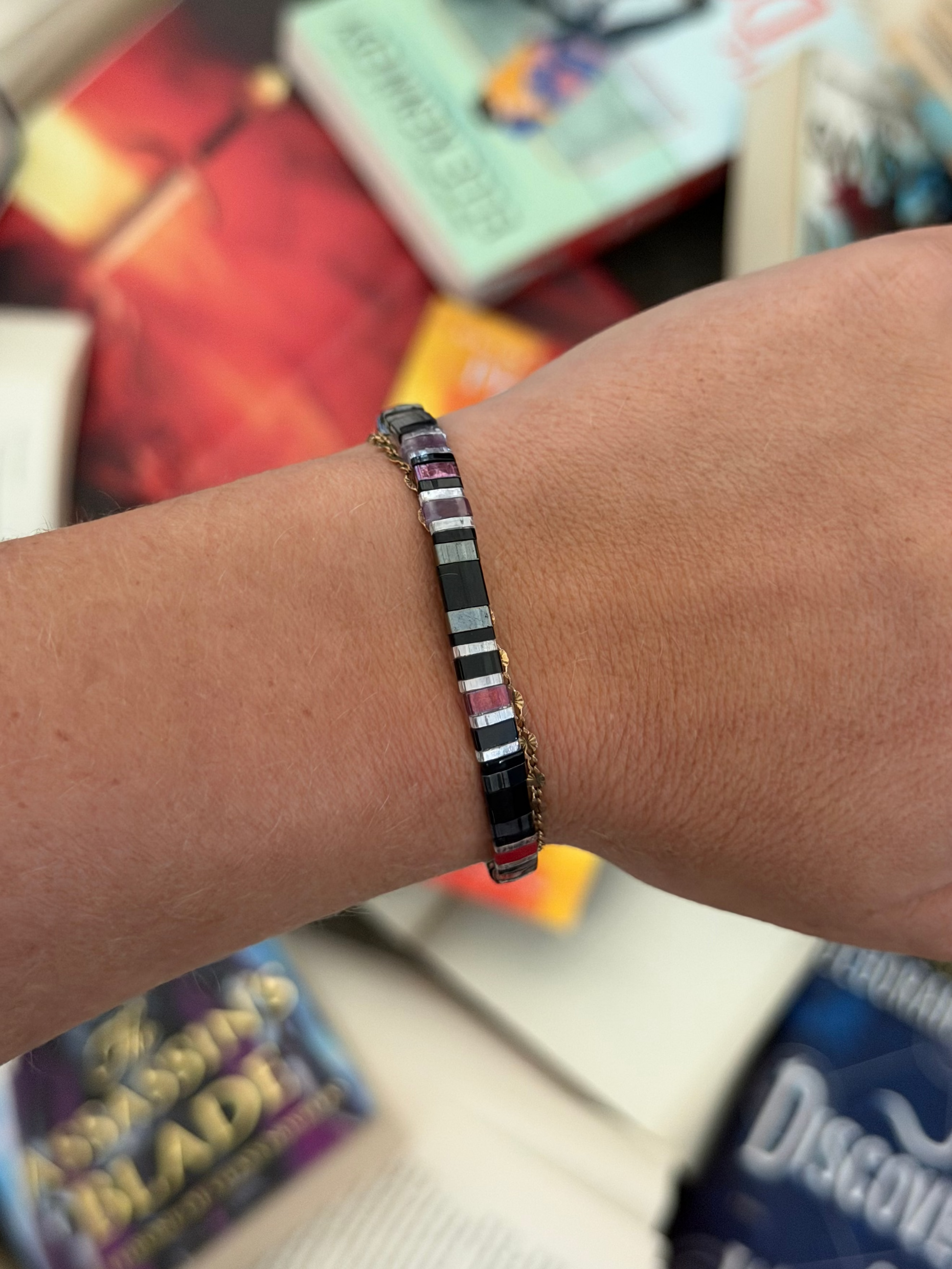 Bat Brothers bracelet showing violet, plum, black, and silver-toned glass beads on a wrist above scattered books.