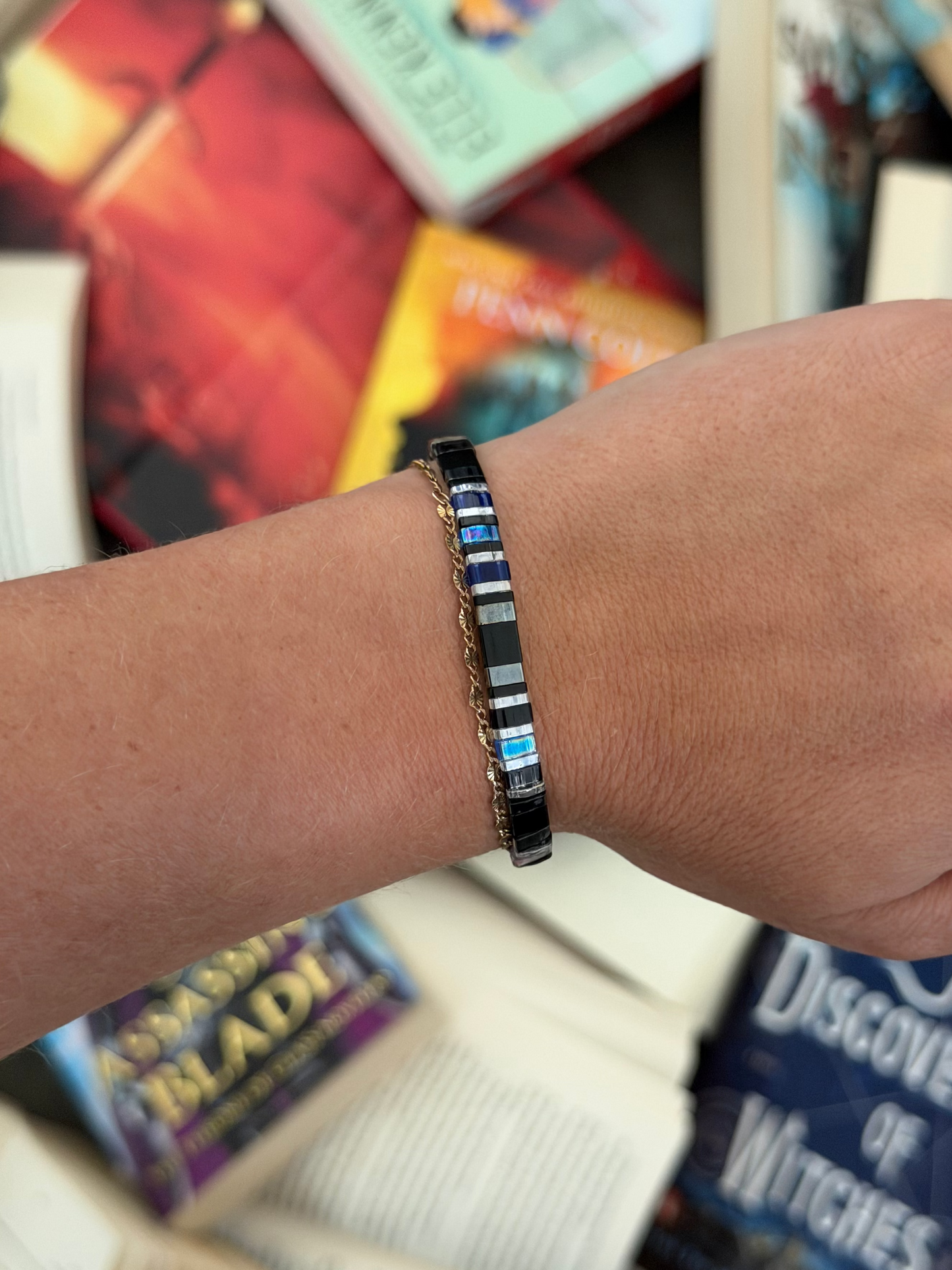Bat Brothers bracelet showing blue, black, and silver-toned glass beads, worn beside a delicate gold bracelet above scattered books.