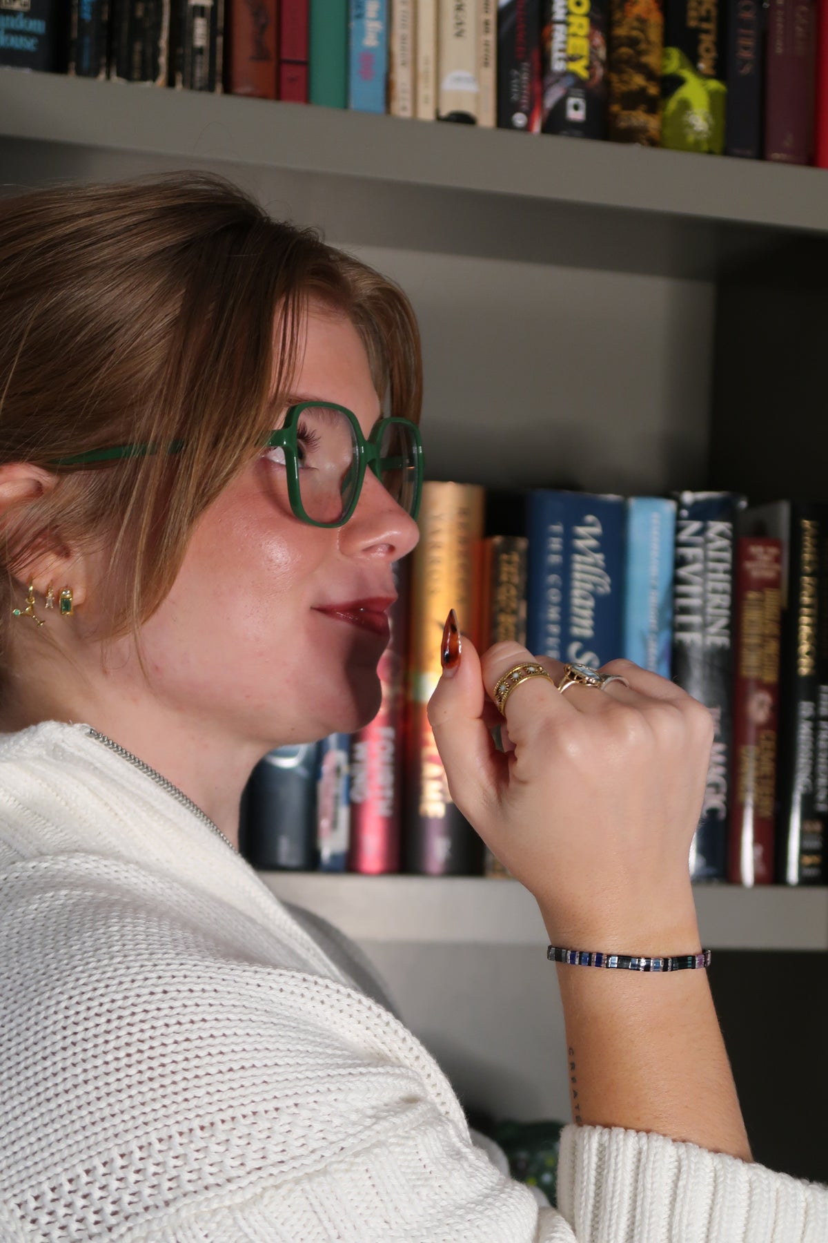 Sarah Baus in green glasses and a cream sweater wearing the Bat Brothers bracelet with her hand raised beside her face in front of bookshelves.