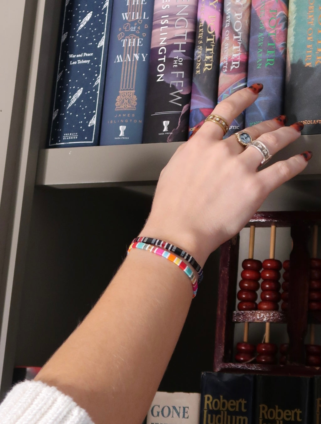 Hand reaching for books on a shelf while wearing the Fantasy Bookshelf Second Edition and Bat Brothers bracelets together.