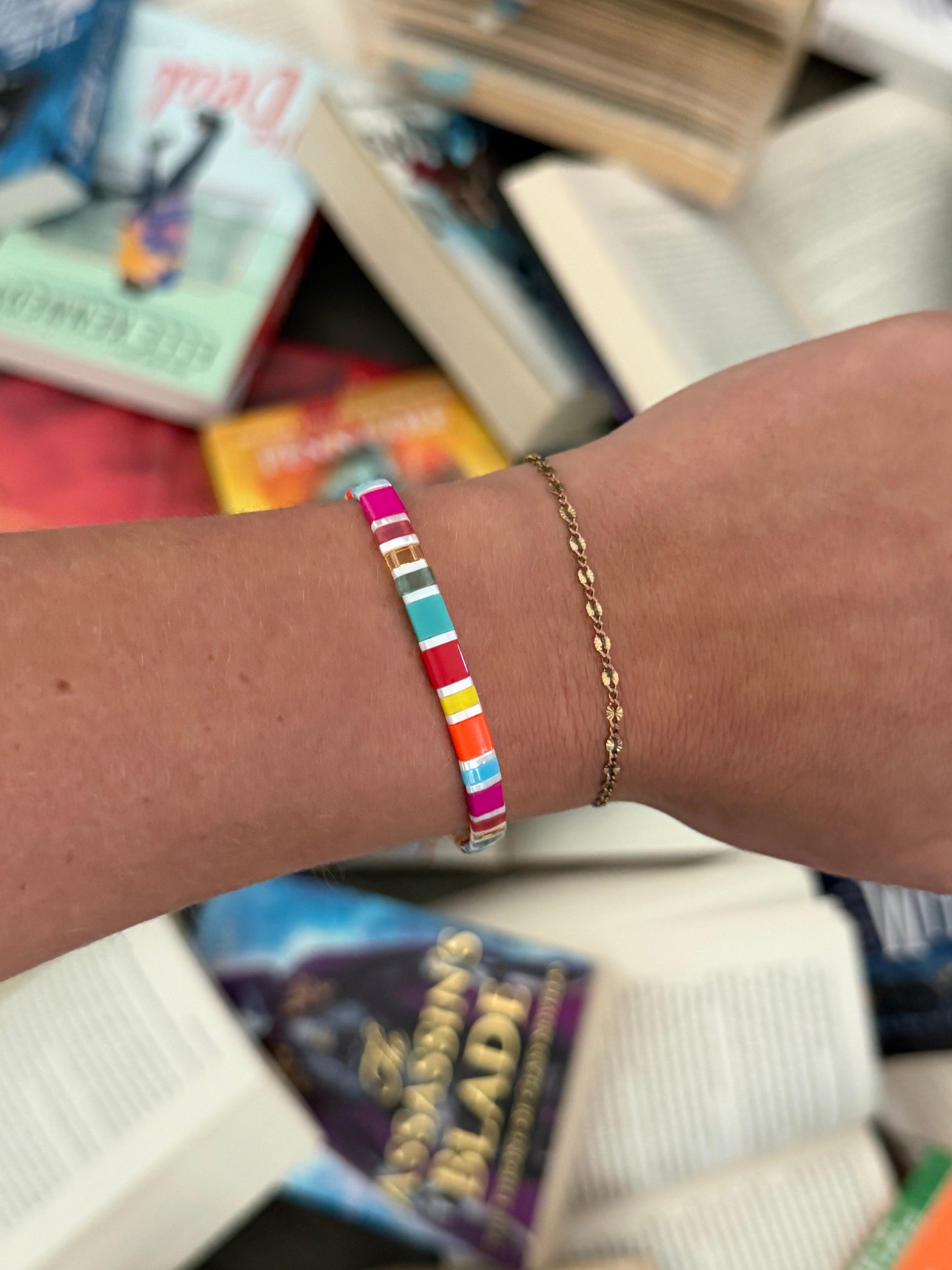 Fantasy Bookshelf Bracelet Second Edition showing colorful rectangular glass beads and white accents, worn beside a delicate gold bracelet above scattered books.