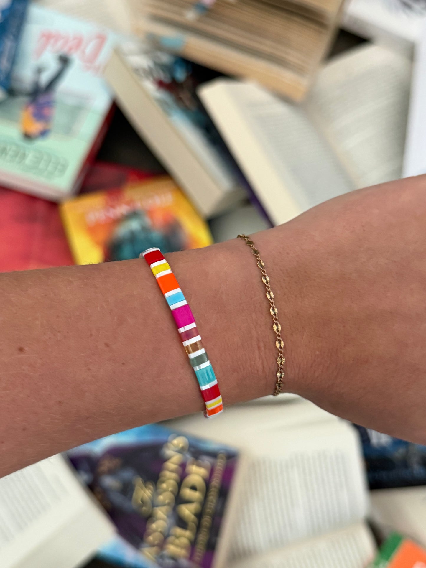 Fantasy Bookshelf Bracelet Second Edition showing colorful tila glass beads and white accents, worn beside a delicate gold bracelet above scattered books.