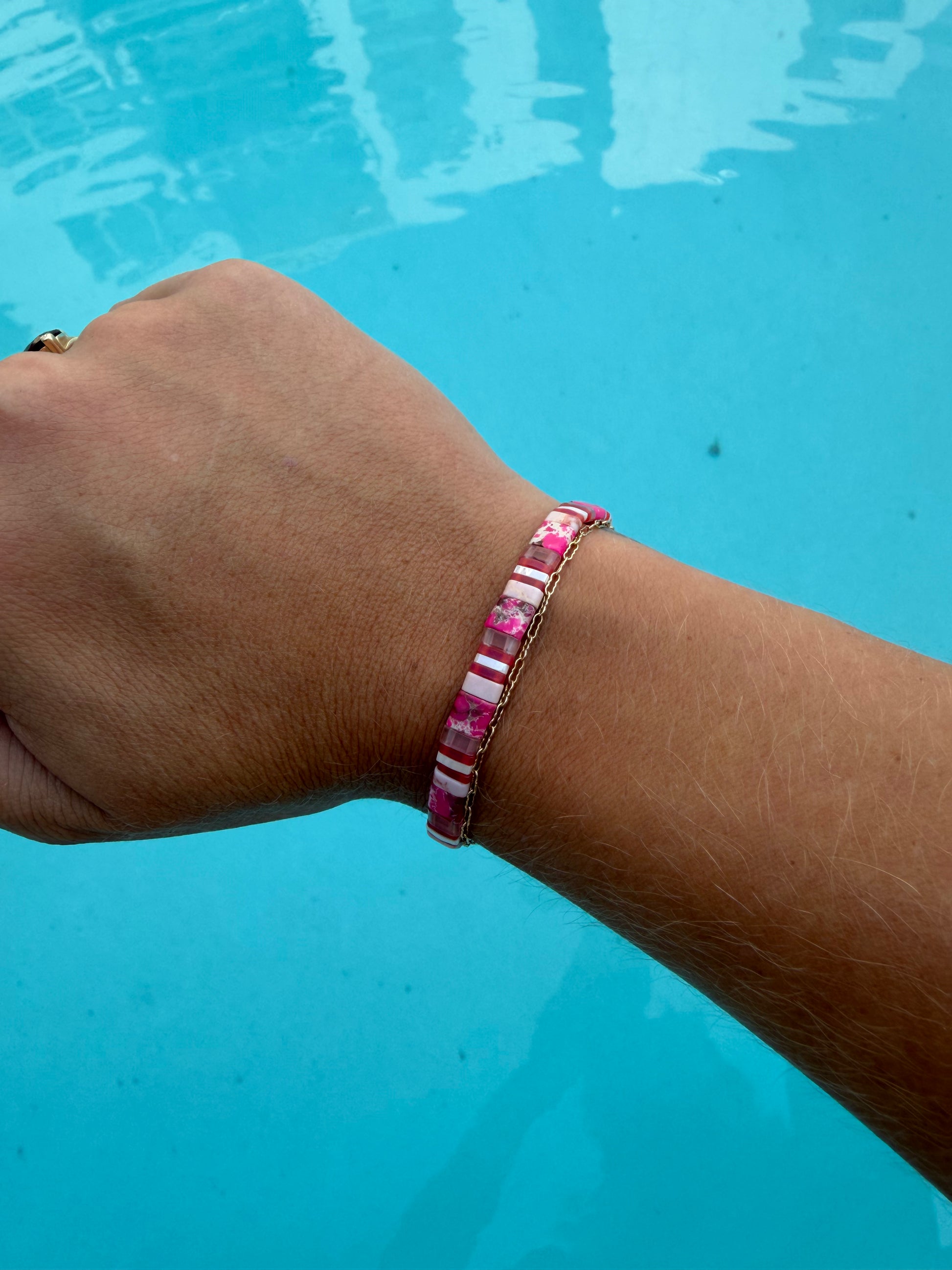 Handmade Pink Sea Star bracelet worn on a wrist beside a gold chain bracelet over blue pool water.