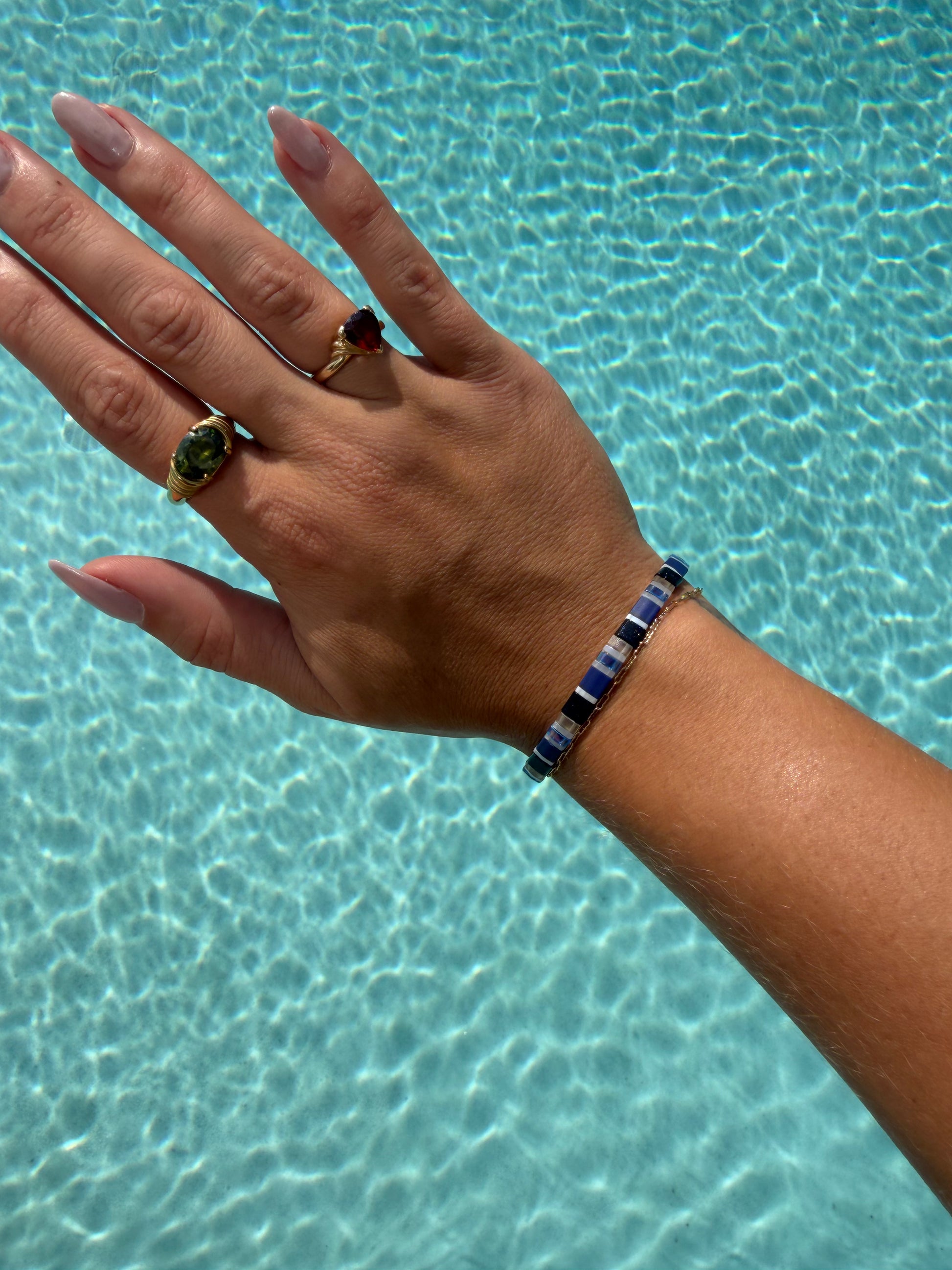 A person's hand and wrist resting over clear blue pool water, wearing a beaded bracelet with alternating dark blue and white beads, along with two rings.