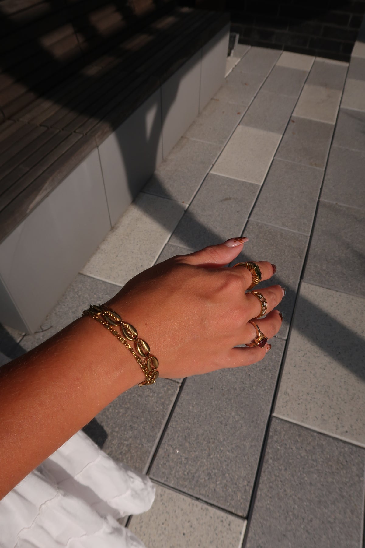 A close-up of a person's hand wearing a gold chain bracelet and matching gold rings, set against a paved outdoor walkway with strong sunlight and shadows.