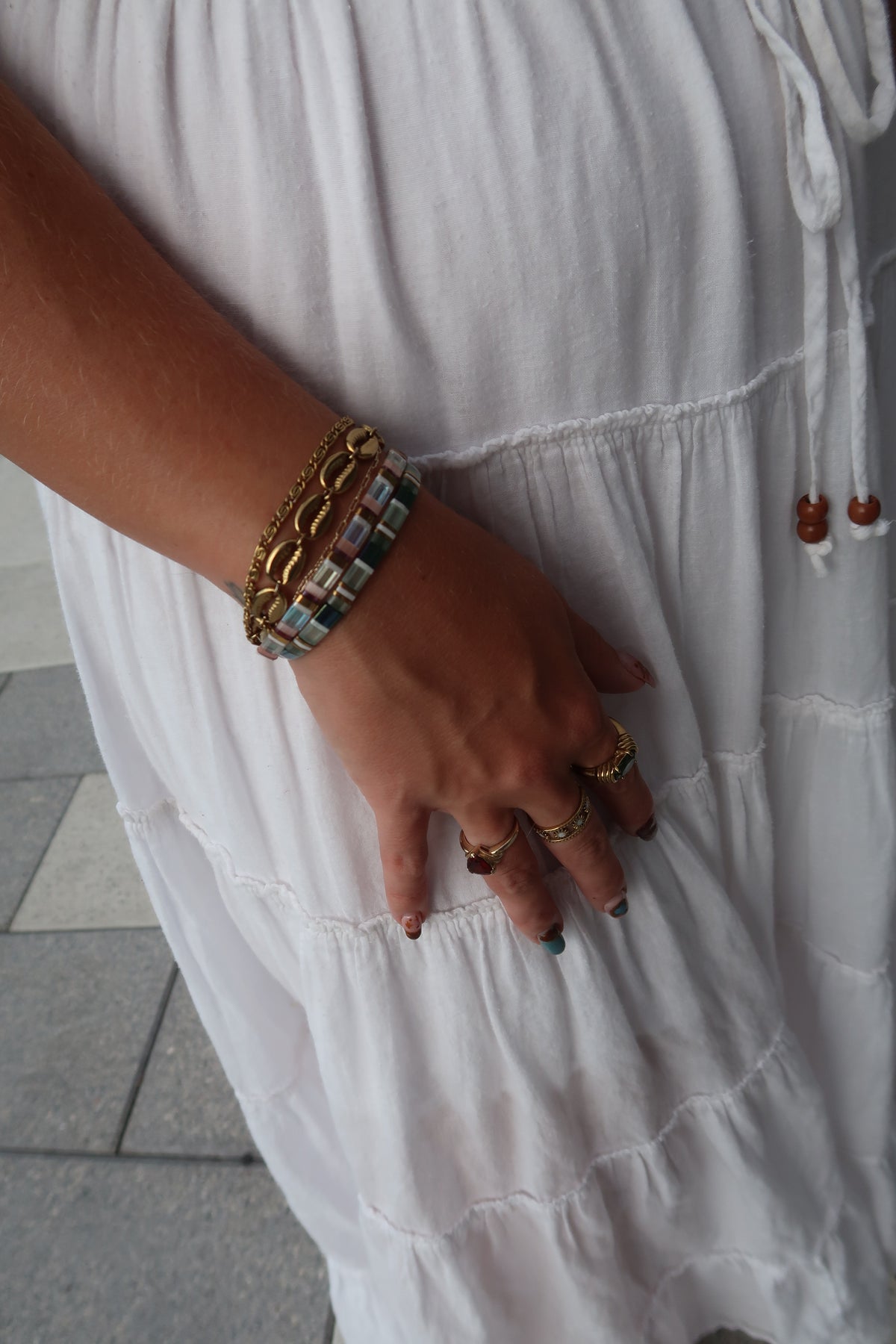 A close-up of a person wearing a white tiered dress, accessorized with a stack of gold chain bracelets: the island cowrie bracelet and whirlpool bracelet paired with the green sea turtle glass-bead bracelet and siren glass-bead bracelet.