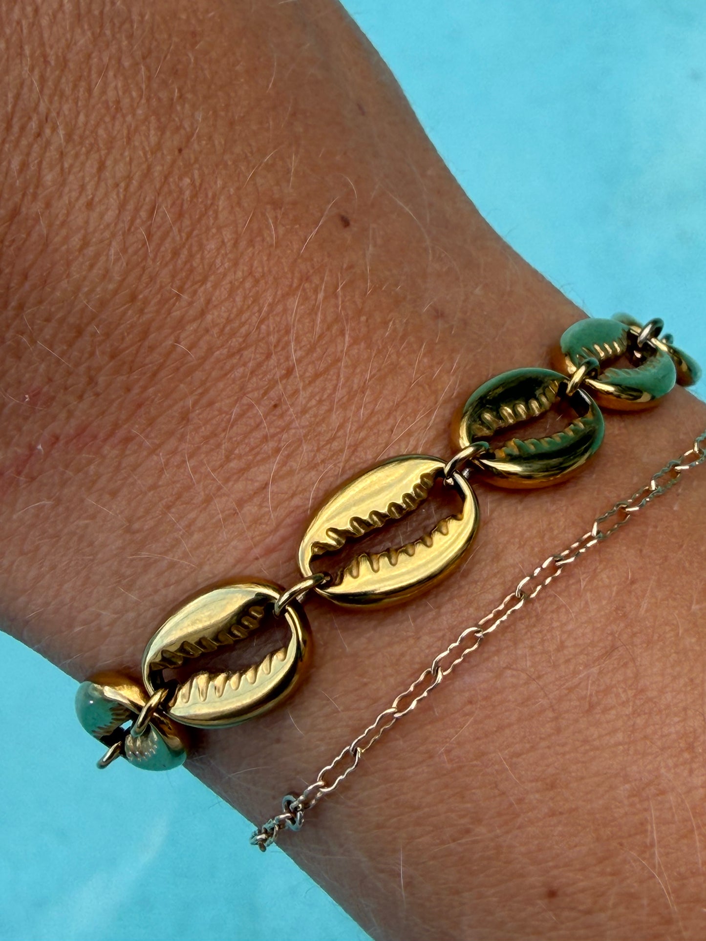 Close-up of the gold Island Cowrie Bracelet layered with a delicate gold chain bracelet by the pool.