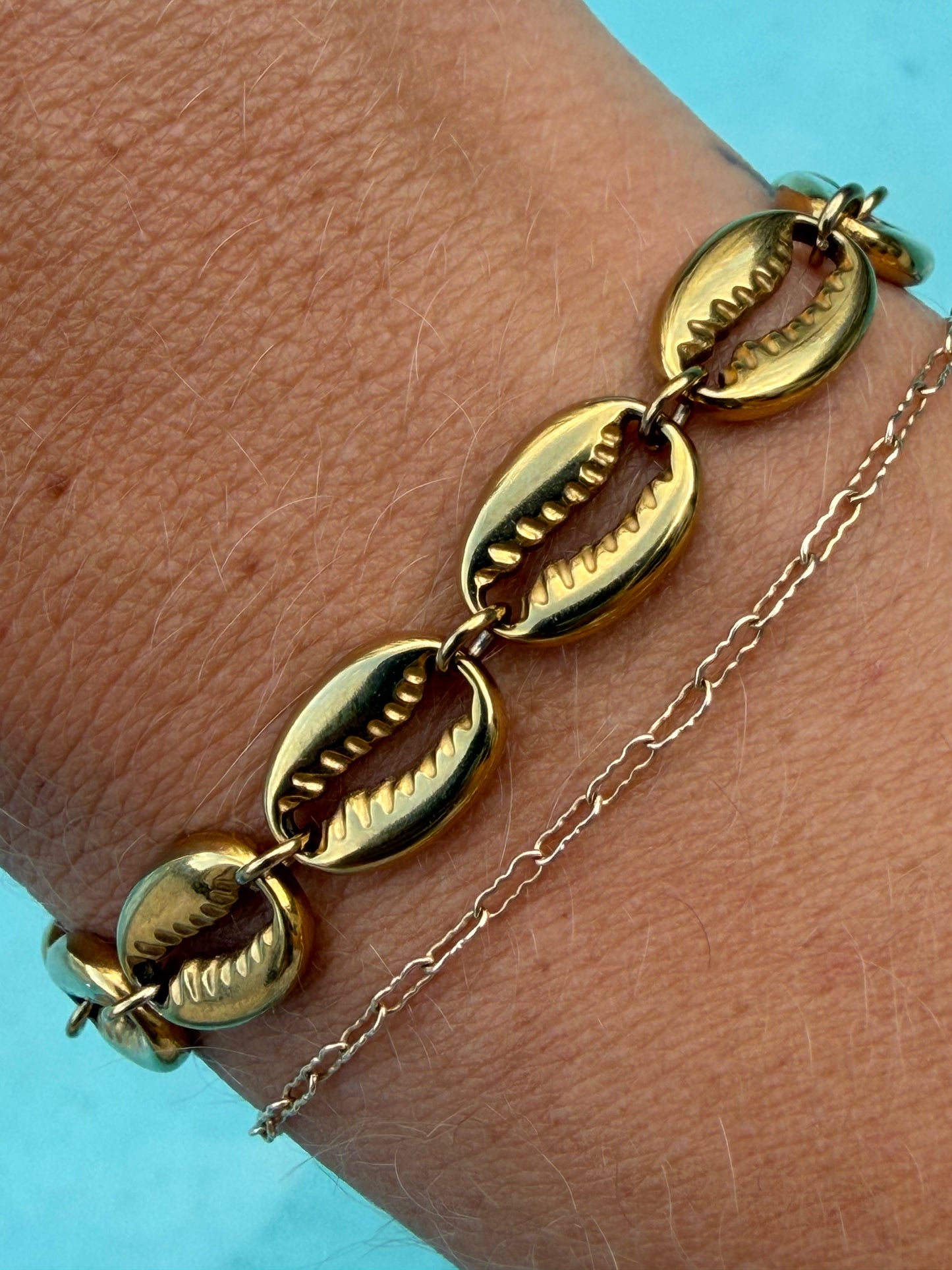 Close-up of the gold Island Cowrie Bracelet layered with a delicate gold chain bracelet by the pool.