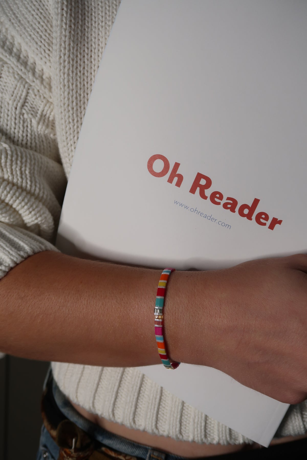 Fantasy Bookshelf Bracelet Second Edition worn on a wrist while holding an Oh Reader magazine against a cream knit sweater.